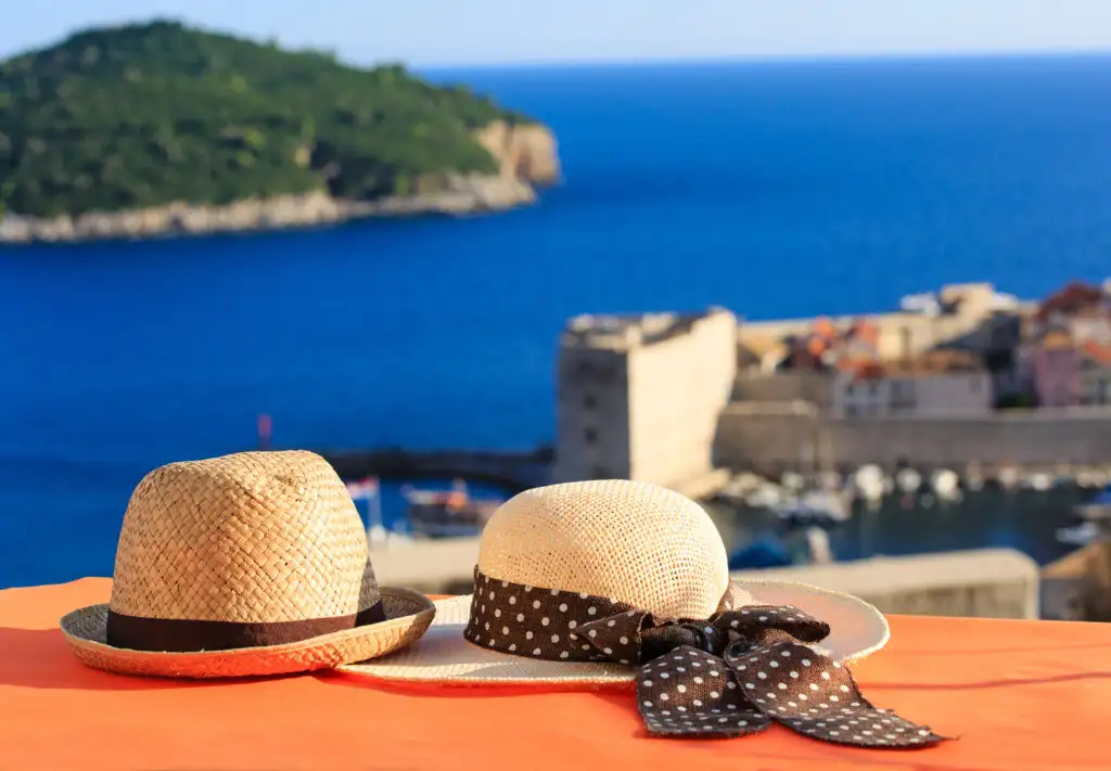 Two hats sitting on a wooden bench overlooking out of focus stone buildings and an ocean bay in the background