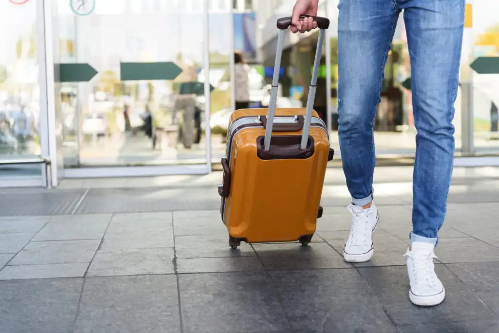 Person wearing jeans and casual white sneakers, pulling rolling luggage into an airport