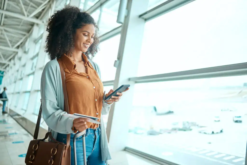 Woman standing in hallway at airport with purse and luggage, looking at her phone