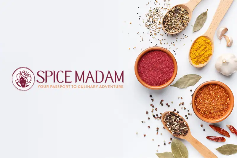 Overhead view of wooden bowls and spoons full of spices alongside the Spice Madam logo