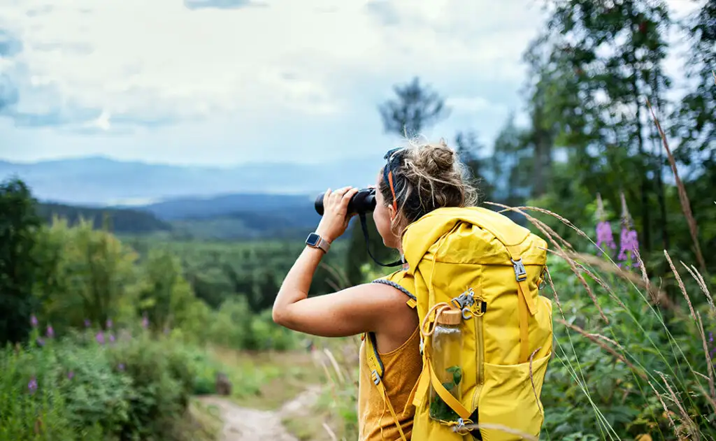 Rear view of woman hiker with backpack on a hiking trip in nature, using binoculars.