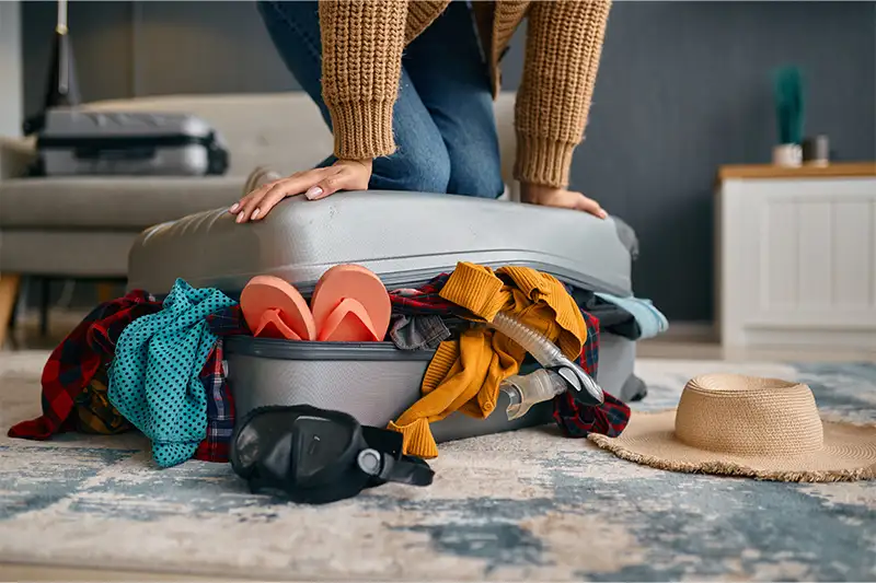 Woman kneeling on top of full overflowing suitcase, trying to close it