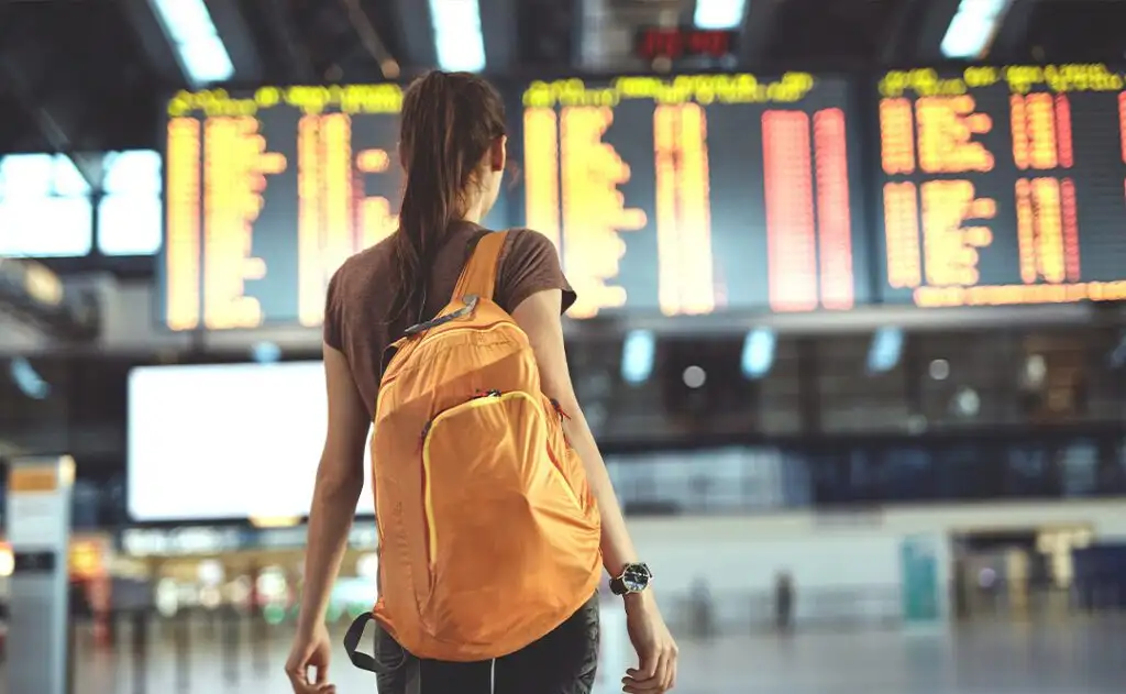 Young woman with small backpack as a hand luggage in international airport looking at the flight information board, checking her flight