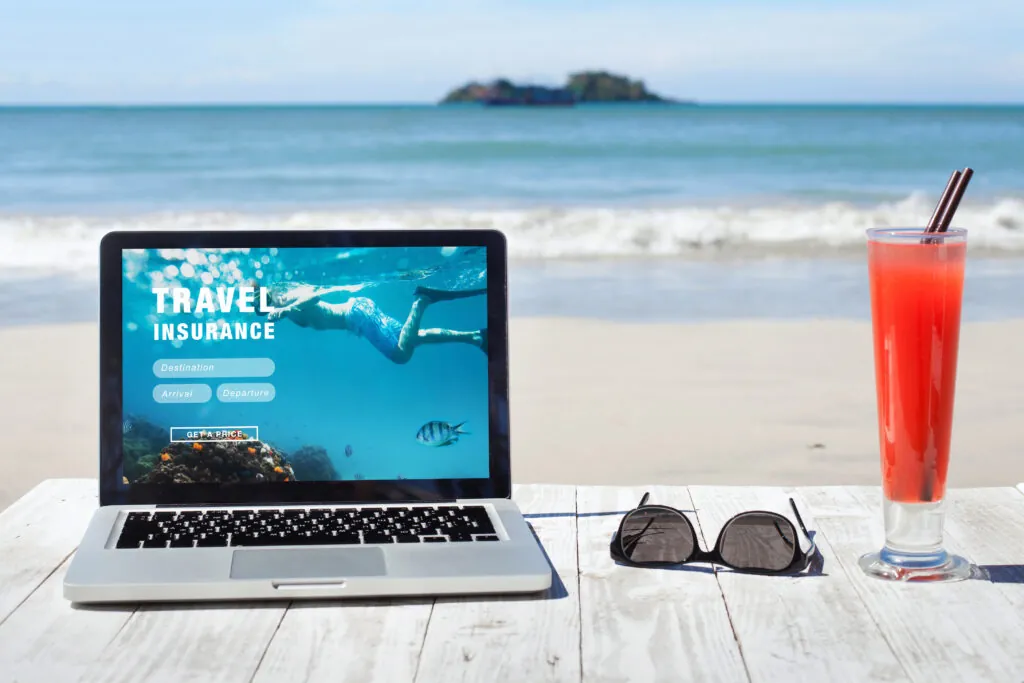 Laptop showing travel insurance site on a table on the beach next to a cocktail in a tall glass and a pair of sunglasses