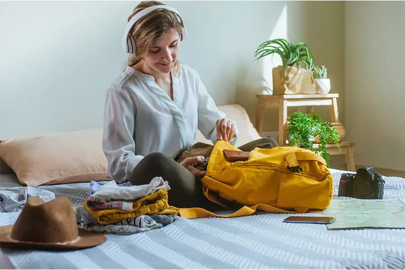 Woman siting on bed and packing travel gear into a yellow backpack
