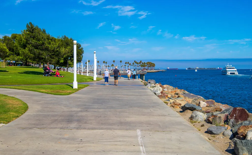 a gorgeous summer landscape in the park with blue ocean water and people relaxing and walking surrounded by lush green trees, grass and plants with blue sky at Shoreline Aquatic Park in Long Beach