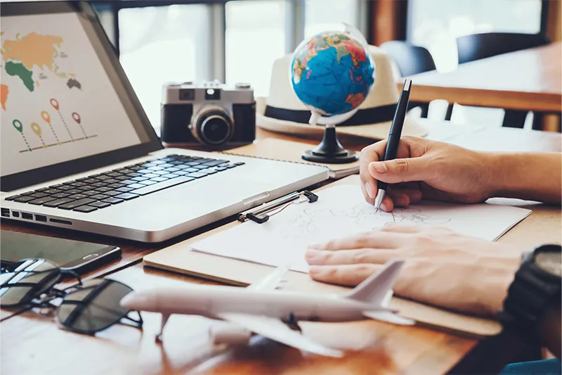 Close up of person writing on a desk surrounded by a globe, camera, model plane, and open laptop showing a world map