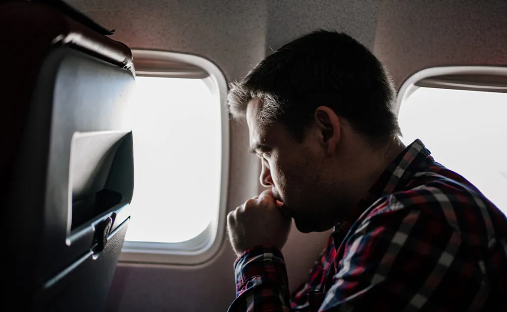 a man in a plaid shirt coughs on the plane in a window seat.