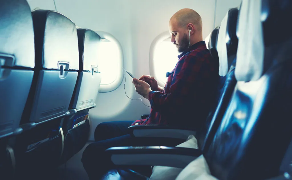 Man is listening to music in headphones and chatting in social network via mobile phone, during his flying in an airplane. Hipster guy is watching video on cell telephone , while is sitting in plane