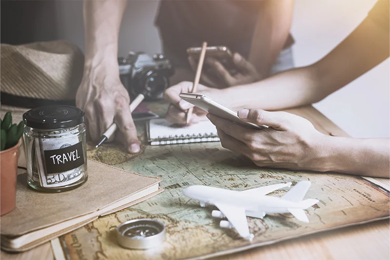 Close up of a couple planning a trip on a map next to a jar full of cash labeled 
